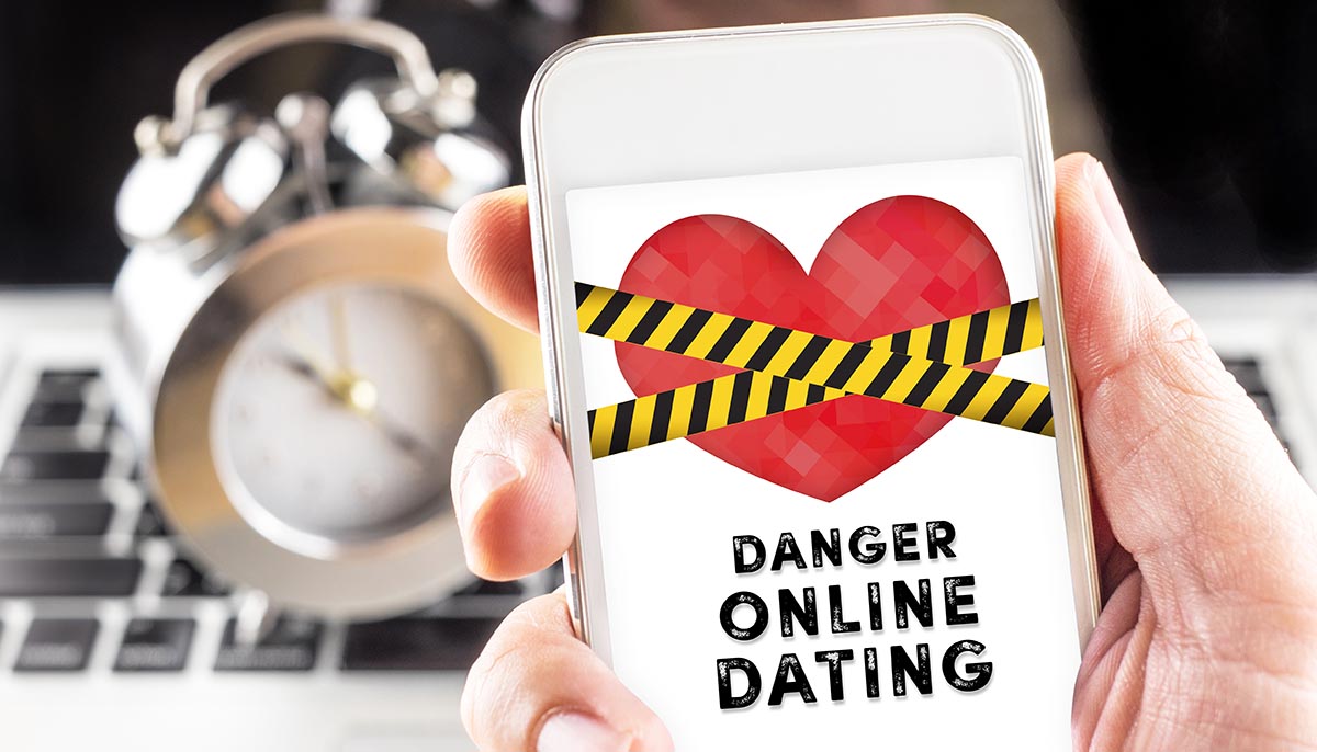 Hand holding mobile with caution tape on heart and Danger online dating on screen with clock and laptop at background, Internet love concept.