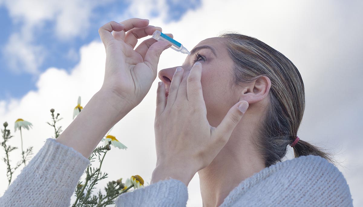 Young woman using eyedropper to treat irritated eyes at flowers meadow